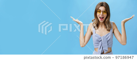 Waist-up shot of charismatic cute young woman in sunglasses raising hands sideways in clueless and unsure gesture shrugging with opened mouth and silly smile being unbothered, posing against blue wall 97186947