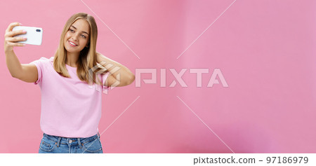 Female beauty blogger taking selfie post new look online. Portrait of charming tanned young woman in t-shirt touching hair gently pulling hand with smartphone near face taking photo over pink wall 97186979