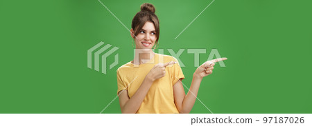 Waist-up shot of charismatic happy and carefree charming woman in yellow t-shirt pointing and looking left enthusiastic and pleased smiling cheerfully posing against green background delighted 97187026