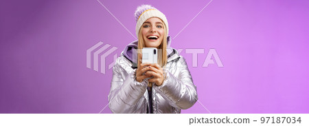 Amused excited attractive blond girlfriend holding smartphone up recording video boyfriend step snowboard first time capturing memories mobile camera standing happily purple background 97187034