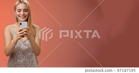 Delighted tender glamour blond woman in silver stylish glittering dress brilliand earrings holding smartphone taking photo friend capture moment celebration nightclub, red background smiling 97187195