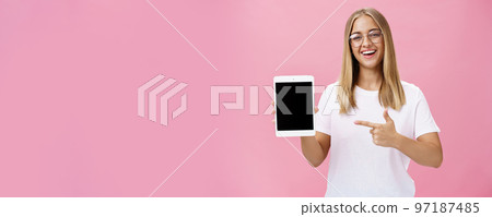 Female freelance programmer proudly showing her app for digital tablet holding gadget pointing at device screen smiling broadly with delighted expression wearing glasses, posing over pink wall 97187485