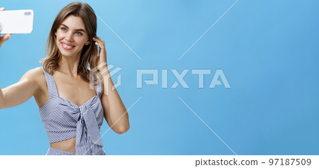 Attractive woman with good self-esteem in stylish matching outfit flicking hair behind ear smiling joyfully at smartphone screen, taking selfie to post in internet and gain followers over blue wall Attractive woman with good self-esteem in stylish matching outfit flicking hair behind ear smiling joyfully at smartphone screen, taking selfie to post in internet and gain followers over blue wall 97187509