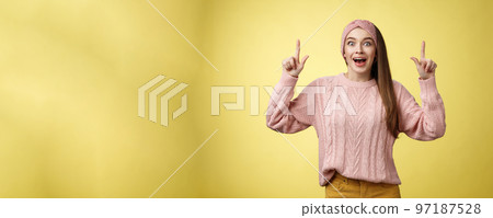 Ecstatic amused positive young 20s woman wearing casual sweater, headband dropping jaw amazed looking astonished, surprised pointing up excited, upbeat of seeing incredible promo over yellow wall 97187528