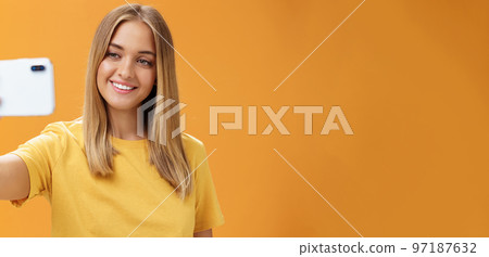Waist-up shto of outgoing and charismatic pretty confident woman with no makeup and tanned skin holding smartphone pulled on distance smiling broadly at cellphone screen while taking selfie 97187632