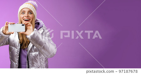 Amused fascinated female in silver jacket head smiling astonished excited look forward holding smartphone recording video taking shots famous person mobile phone camera, purple background 97187678