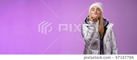 Romantic tender gentle cute girlfriend blond hair wearing hat stylish silver jacket close eyes folding lips muah hold hand near lips blow air kiss passionately feel love sympathy, purple background 97187786