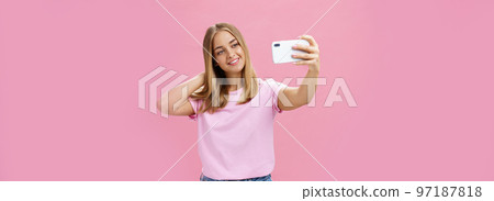 Female beauty blogger taking selfie post new look online. Portrait of charming tanned young woman in t-shirt touching hair gently pulling hand with smartphone near face taking photo over pink wall 97187818