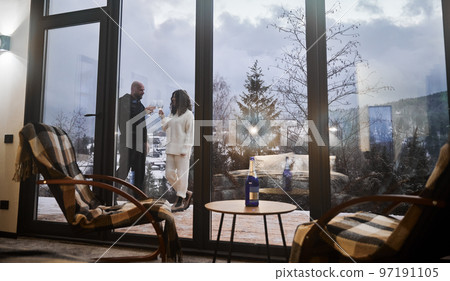 Beautiful view of man and woman toasting with wine behind panoramic window of scandinavian house barnhouse. Interior of wooden house room with chairs and couple in love outside building. Beautiful view of man and woman toasting with wine behind panoramic window of scandinavian house barnhouse. Interior of wooden house room with chairs and couple in love outside building. 97191105