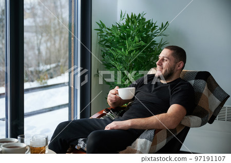 Young man enjoying weekends inside contemporary barn house. Happy male tourist sitting on chair, holding cup of tea, enjoying view of mountain landscape through panoramic windows in new cottage. 97191107