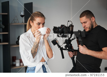 Videographer man shooting footage with female model, using camera mounted on gimbal stabilizer equipment indoors. Young woman with tattoos on hands posing and drinking hot tea. 97191153
