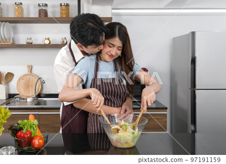 Asian couple spend time together in the kitchen. Young man hugs and kisses his girlfriend's cheek from behind while the woman mix the vegetables in a salad bowl together with a wooden ladle. Asian couple spend time together in the kitchen. Young man hugs and kisses his girlfriend's cheek from behind while the woman mix the vegetables in a salad bowl together with a wooden ladle. 97191159