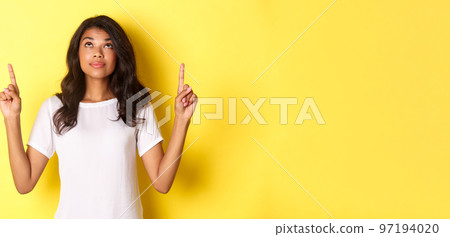 Portrait of attractive african-american female model, wearing white t-shirt, looking and pointing fingers up with curious smile, showing promo, standing over yellow background Portrait of attractive african-american female model, wearing white t-shirt, looking and pointing fingers up with curious smile, showing promo, standing over yellow background 97194020