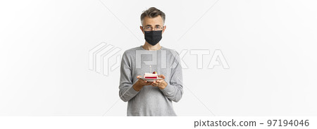Concept of coronavirus, quarantine and holidays. Image of sad middle-aged man in black medical mask and glasses, holding birthday cake with lit candle, feel upset during b-day celebration Concept of coronavirus, quarantine and holidays. Image of sad middle-aged man in black medical mask and glasses, holding birthday cake with lit candle, feel upset during b-day celebration 97194046