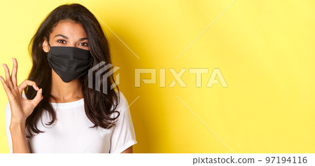 Concept of coronavirus, pandemic and lifestyle. Close-up of pretty african-american woman in black face mask, showing okay sign in approval, praise good work, yellow background Concept of coronavirus, pandemic and lifestyle. Close-up of pretty african-american woman in black face mask, showing okay sign in approval, praise good work, yellow background 97194116