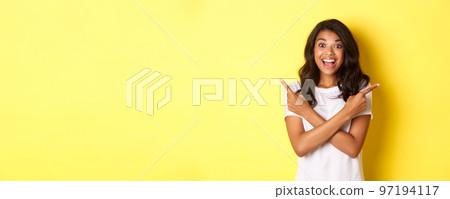 Portrait of happy and amazed african american woman, showing two promo offers, pointing fingers sideways and smiling excited, standing over yellow background Portrait of happy and amazed african american woman, showing two promo offers, pointing fingers sideways and smiling excited, standing over yellow background 97194117