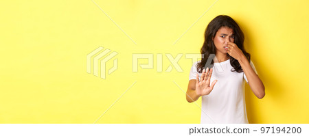 Young african-american woman in white t-shirt, shut her nose, looking disgusted and asking to stay away, dislike bad smell, standing over yellow background Young african-american woman in white t-shirt, shut her nose, looking disgusted and asking to stay away, dislike bad smell, standing over yellow background 97194200