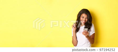 Image of attractive african american girl in white t-shirt, messaging on smartphone, looking at mobile phone and smiling, standing over yellow background Image of attractive african american girl in white t-shirt, messaging on smartphone, looking at mobile phone and smiling, standing over yellow background 97194252