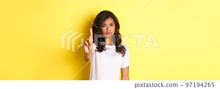 Image of disappointed african-american girl telling no, shaking finger to prohibit or stop someone, disagree with person, standing over yellow background 97194265