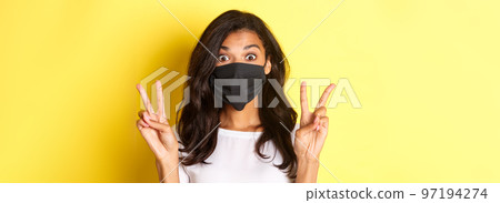 Concept of coronavirus, pandemic and lifestyle. Close-up of silly african-ameircan girl in black face mask, showing peace signs and standing over yellow background Concept of coronavirus, pandemic and lifestyle. Close-up of silly african-ameircan girl in black face mask, showing peace signs and standing over yellow background 97194274