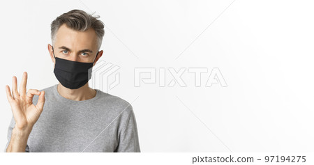 Concept of covid-19, social distancing and quarantine. Close-up of confident middle-aged man, wearing black medical mask, showing okay sign and praising something good Concept of covid-19, social distancing and quarantine. Close-up of confident middle-aged man, wearing black medical mask, showing okay sign and praising something good 97194275