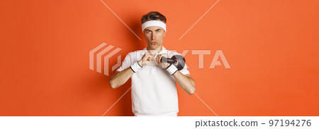 Concept of workout, gym and lifestyle. Close-up of determined middle-aged fitness guy, doing sport exercises with kettlebell, standing over orange background Concept of workout, gym and lifestyle. Close-up of determined middle-aged fitness guy, doing sport exercises with kettlebell, standing over orange background 97194276