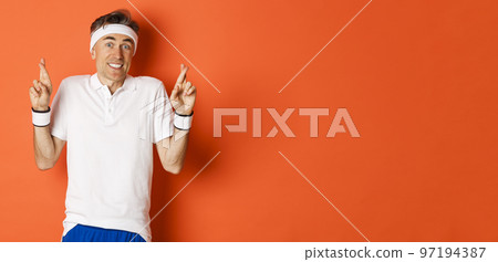 Concept of sport, fitness and lifestyle. Portrait of hopeful middle-aged male athlete, crossing fingers for good luck and making wish, standing over orange background Concept of sport, fitness and lifestyle. Portrait of hopeful middle-aged male athlete, crossing fingers for good luck and making wish, standing over orange background 97194387