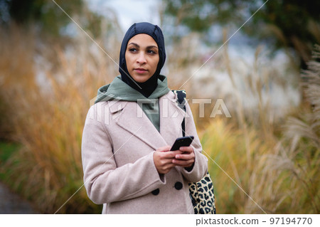 Portrait of young muslim woman in coat, standing outdoor in city park, scrolling smartphone. Portrait of young muslim woman in coat, standing outdoor in city park, scrolling smartphone. 97194770