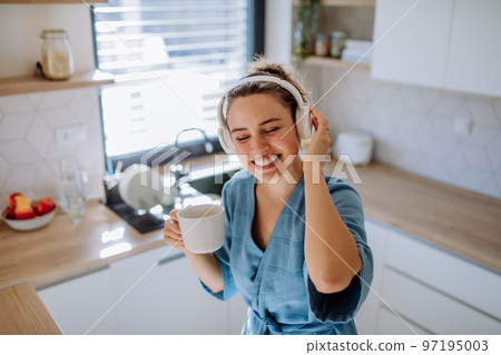 Young woman listening music and enjoying cup of coffee at morning, in her kitchen. 97195003