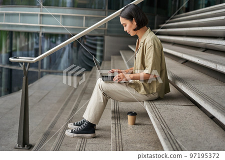 Profile portrait of young asian woman with laptop, girl student sits on stairs outside building and types on computer, drinks takeaway coffee 97195372
