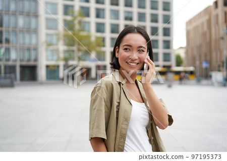Modern young asian girl talks on mobile phone, uses telephone on city street. Woman smiling while calling someone on smartphone Modern young asian girl talks on mobile phone, uses telephone on city street. Woman smiling while calling someone on smartphone 97195373