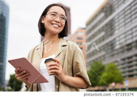 Portrait of happy young woman in glasses, standing on street with cup of coffee and tablet 97195374