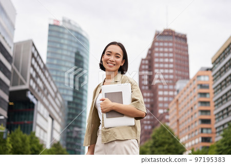 Portrait of young asian woman, looking happy and confident, going to work or university, city skyscrappers behind her, holding laptop and notebook Portrait of young asian woman, looking happy and confident, going to work or university, city skyscrappers behind her, holding laptop and notebook 97195383