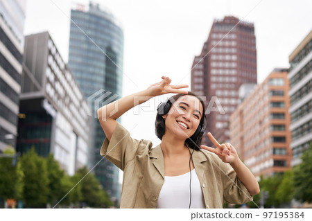 Happy asian woman in headphones, listening music and dancing on street of city centre, smiling with hands up Happy asian woman in headphones, listening music and dancing on street of city centre, smiling with hands up 97195384