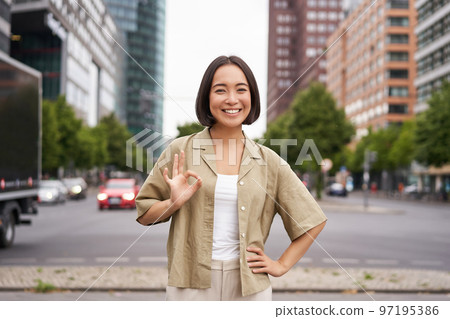 Enthusiastic city girl, shows okay gesture in approval, looking upbeat, say yes, approves and agrees, stands on street Enthusiastic city girl, shows okay gesture in approval, looking upbeat, say yes, approves and agrees, stands on street 97195386