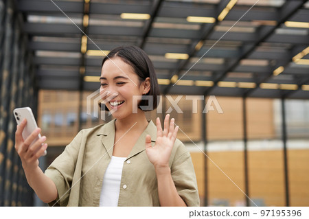 Happy asian girl waves at her smartphone camera, says hello to friend on video chat, calling someone, standing on street Happy asian girl waves at her smartphone camera, says hello to friend on video chat, calling someone, standing on street 97195396