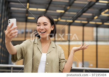Happy asian girl shows something behind her during video call, demonstrating smth, standing on street 97195404