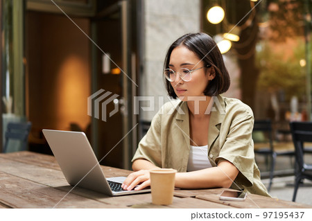 Young woman working in a cafe, using laptop and drinking coffee. Asian girl student with computer studying remotely, sitting on bench near shop 97195437