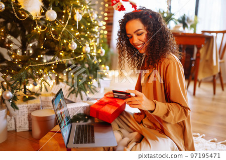 Smiling African American woman using laptop, sitting near Christmas tree with card, shopping online 97195471