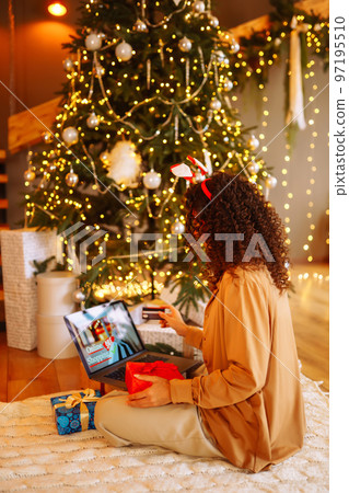 Smiling African American woman using laptop, sitting near Christmas tree with card, shopping online Smiling African American woman using laptop, sitting near Christmas tree with card, shopping online 97195510