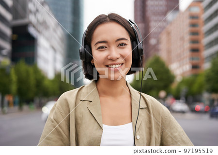 Portrait of smiling asian woman in headphones, standing in city centre on street, looking happy, listening to music Portrait of smiling asian woman in headphones, standing in city centre on street, looking happy, listening to music 97195519