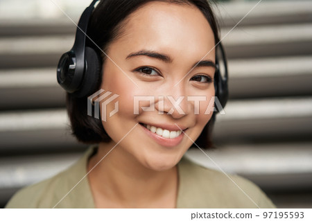 Close up portrait of smiling asian girl in headphones, listens to music outdoors, looking happy. 97195593
