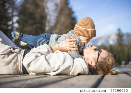 Happy family. Young mother playing with her baby boy infant oudoors on sunny autumn day. Portrait of mom and little son on wooden platform by lake. Positive human emotions, feelings, joy Happy family. Young mother playing with her baby boy infant oudoors on sunny autumn day. Portrait of mom and little son on wooden platform by lake. Positive human emotions, feelings, joy 97196010