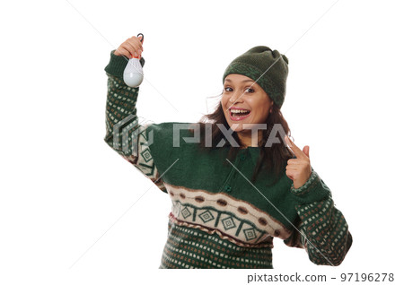 Happy Latin American woman in warm clothes, smiling a cheerful toothy smile looking at camera, pointing at a flashlight bulb. Solution in case of lack or interruption of electricity, white background 97196278