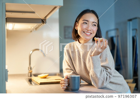 Beautiful young asian woman eating toast on her kitchen, drinking coffee and smiling Beautiful young asian woman eating toast on her kitchen, drinking coffee and smiling 97196307