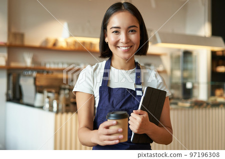 Smiling barista, cafe waitress giving out takeaway coffee order, prepared cappuccino for takeout, standing in blue apron with tablet Smiling barista, cafe waitress giving out takeaway coffee order, prepared cappuccino for takeout, standing in blue apron with tablet 97196308