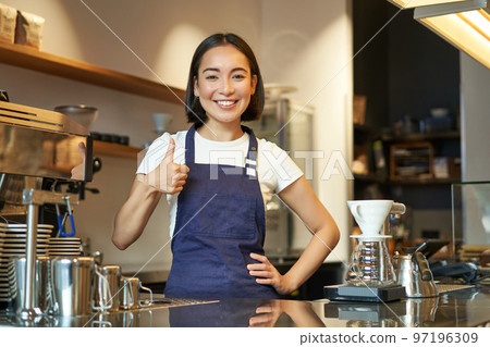 Smiling asian barista, girl in cafe uniform, shows okay sign, recommends their coffee, approve new flavours, standing in apron Smiling asian barista, girl in cafe uniform, shows okay sign, recommends their coffee, approve new flavours, standing in apron 97196309