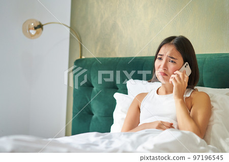 Portrait of concerned asian woman holding mobile phone, receive bad phone call, looking worried and upset, having difficult telephone conversation while lying in bed 97196545