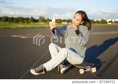 Beautiful korean girl takes selfie on smartphone, takes photo with her skateboard, enoys sunny day outdoors 97196552
