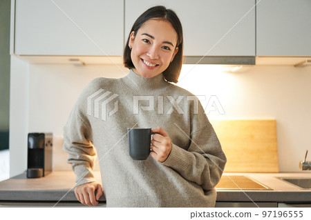 Portrait of smiling asian woman standing on her kitchen, drinking coffee and looking at camera, concept of cozy and comfortable home 97196555
